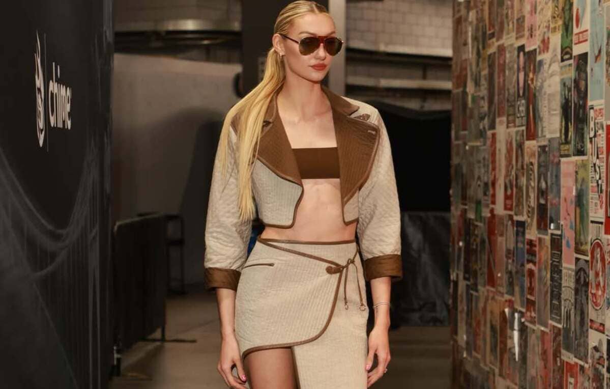 Model walking backstage in a beige and brown two-piece outfit with a cropped jacket and wrap skirt, wearing sunglasses.