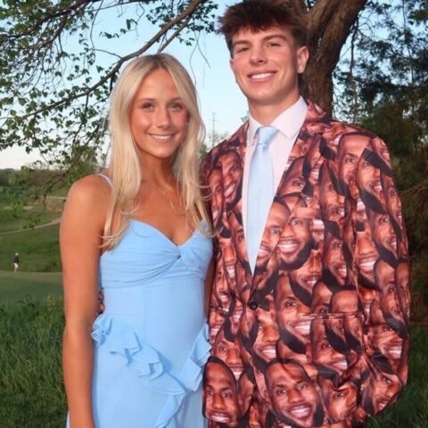 Smiling couple posing outdoors: woman in a light blue dress and man wearing a face‑print suit jacket, standing among trees and grass.