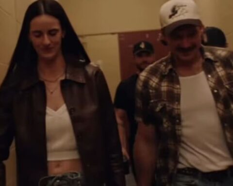 Woman in a brown leather jacket and white crop top walking beside a man in a plaid shirt and white tee down a hallway, both smiling slightly.