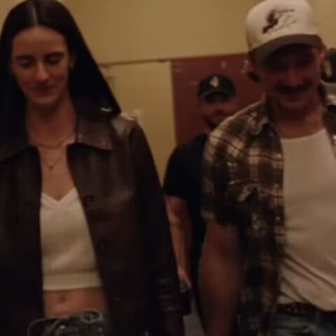 Woman in a brown leather jacket and white crop top walking beside a man in a plaid shirt and white tee down a hallway, both smiling slightly.