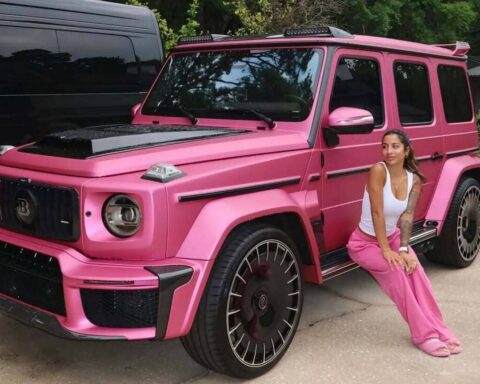 Bright pink Mercedes G-Wagon parked on a driveway, with a woman in a white tank top and pink pants sitting beside it