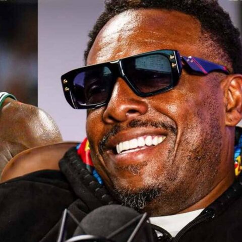 Close-up of a man smiling, wearing oversized black sunglasses, with a microphone in front.