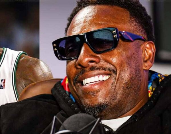 Close-up of a man smiling, wearing oversized black sunglasses, with a microphone in front.