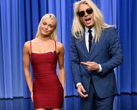 Two blond presenters on a TV stage with blue curtains; woman in a red dress smiles, man in a blue suit and sunglasses points at her.