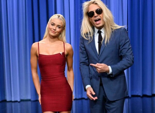 Two blond presenters on a TV stage with blue curtains; woman in a red dress smiles, man in a blue suit and sunglasses points at her.
