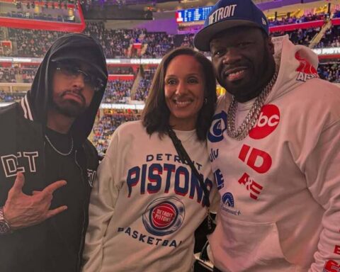 Three people posing in a basketball arena; two wear Detroit Pistons gear and crowd in the background symmetry of bleachers visible at top left and right.