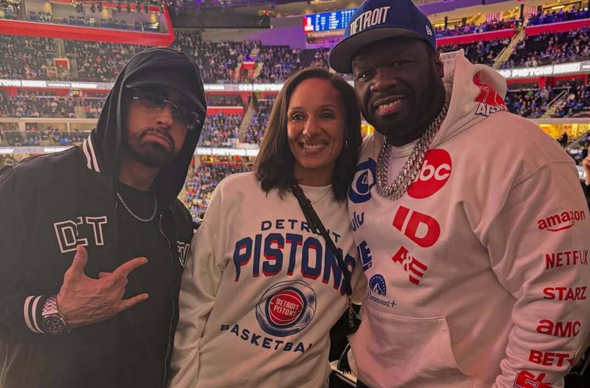 Three people posing in a basketball arena; two wear Detroit Pistons gear and crowd in the background symmetry of bleachers visible at top left and right.