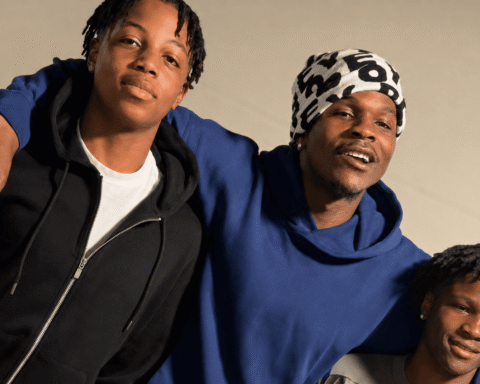 Three friends pose with arms around each other, smiling at the camera; casual hoodies and a patterned headwrap present they stand against a beige backdrop.