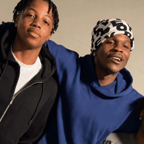 Three friends pose with arms around each other, smiling at the camera; casual hoodies and a patterned headwrap present they stand against a beige backdrop.