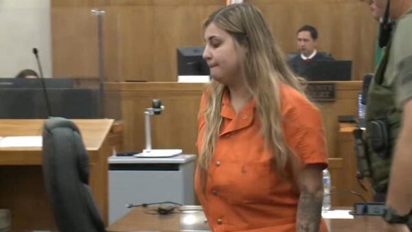 Defendant in an orange jumpsuit walks through a courtroom; a judge sits at the bench in the background.