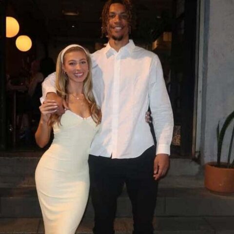Couple posing arm-in-arm outside a doorway at night; woman in a white bodycon dress, man in a white button-down shirt and black pants.