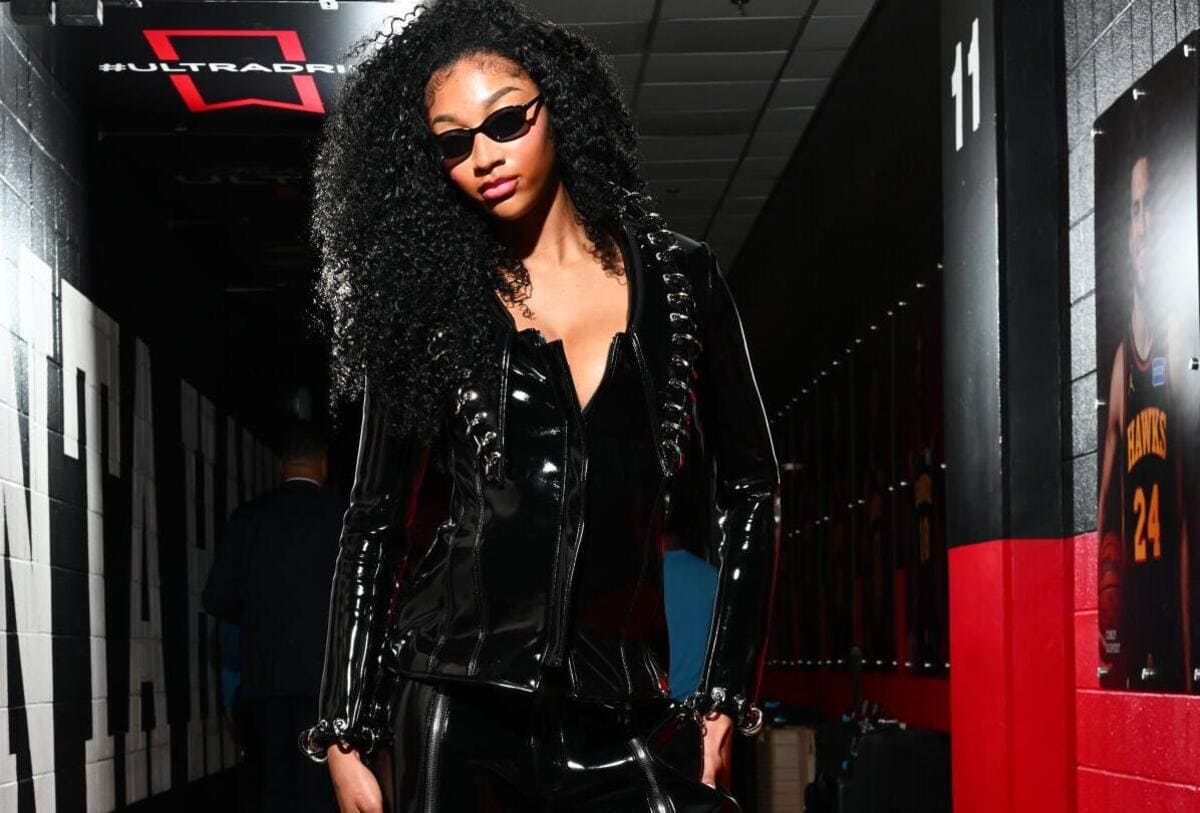 Woman in a black shiny leather outfit and sunglasses walking through a dim backstage hallway.
