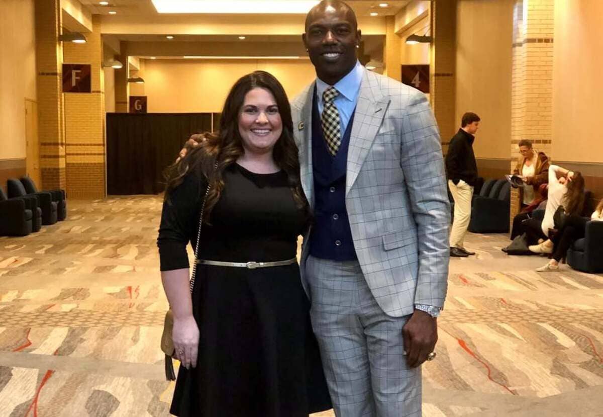 Smiling man in a light gray plaid suit poses with a woman in a black dress inside a conference hallway, both looking at the camera.