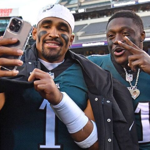 Two football players in teal jerseys take a postgame selfie on the field, one flashing a peace sign toward the camera while smiling.