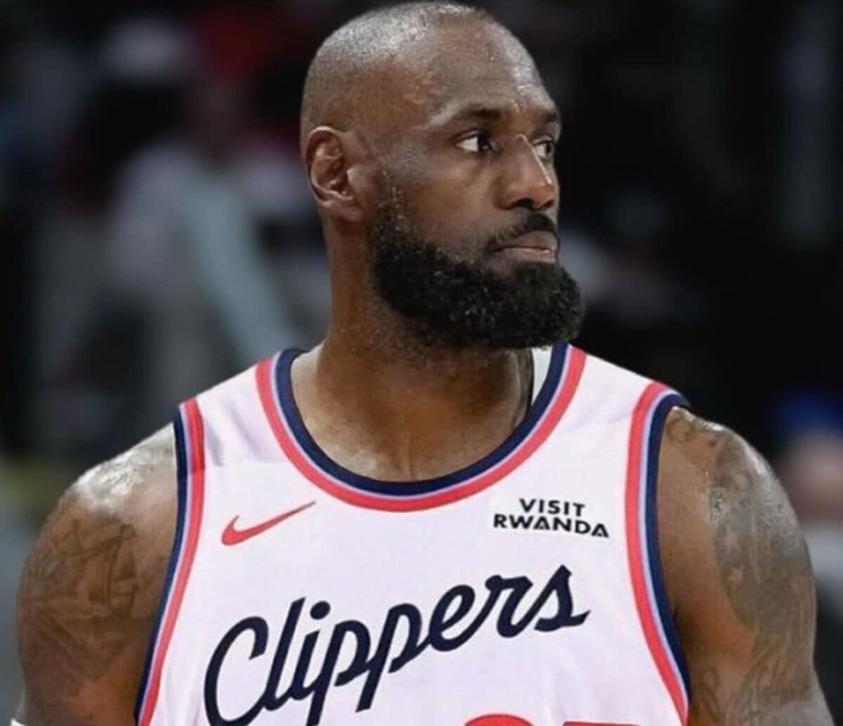 Basketball player in a white Clippers jersey with red and blue trim, looking to the right with a full beard.