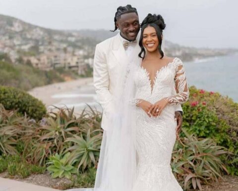 Smiling bride in a lace white wedding gown and groom in a white suit pose together outdoors by the coast with a garden backdrop.