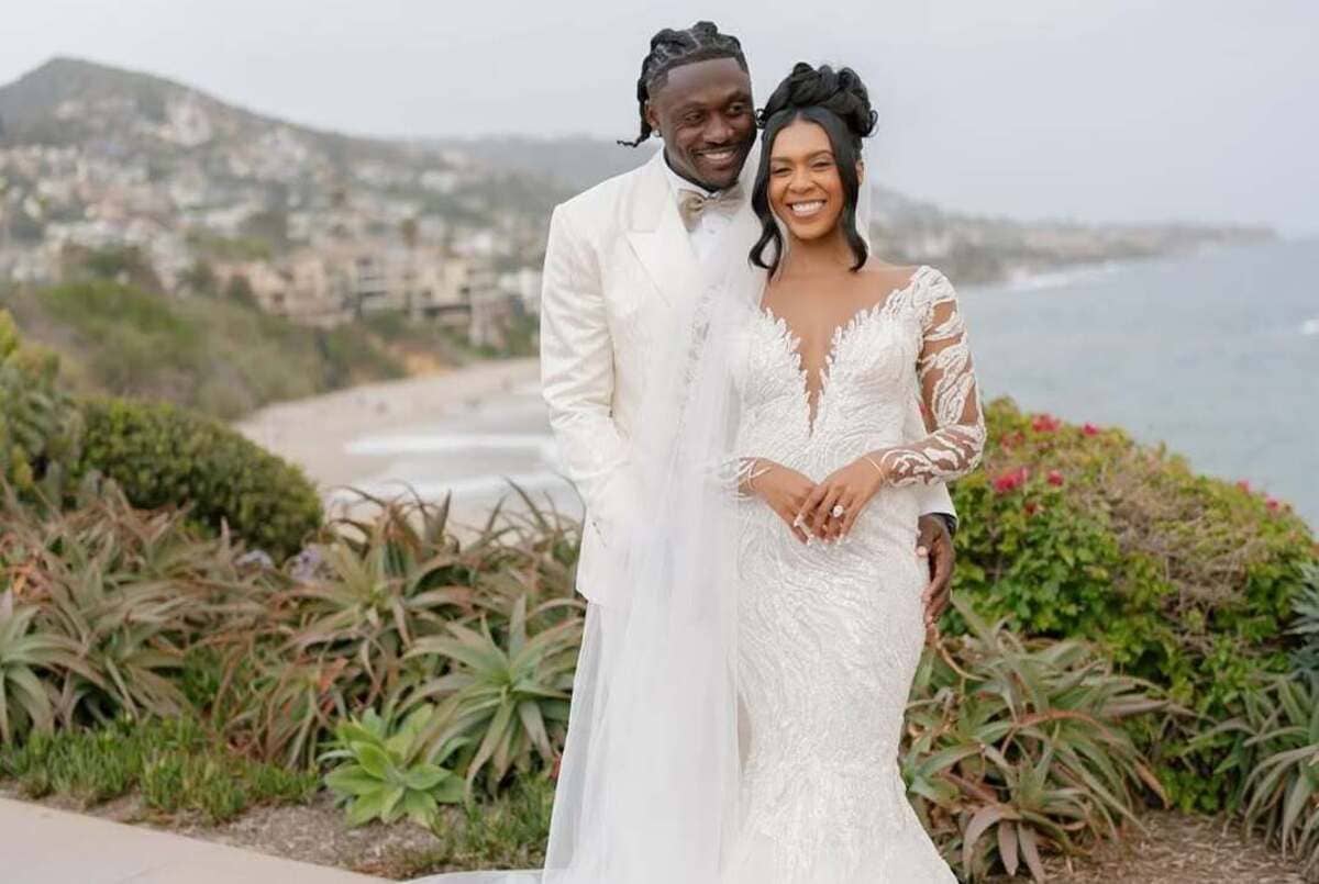 Smiling bride in a lace white wedding gown and groom in a white suit pose together outdoors by the coast with a garden backdrop.