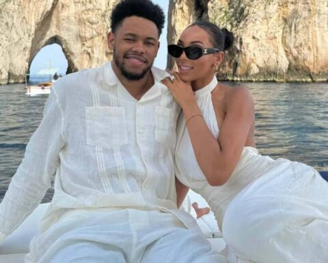 Couple in white outfits sit close on a boat, smiling at the camera with a rocky arch and blue water in the background.