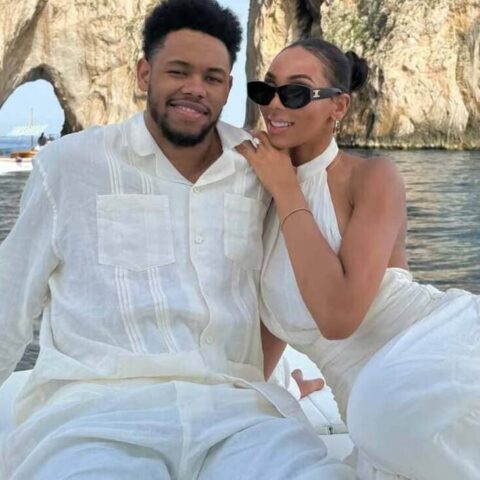 Couple in white outfits sit close on a boat, smiling at the camera with a rocky arch and blue water in the background.