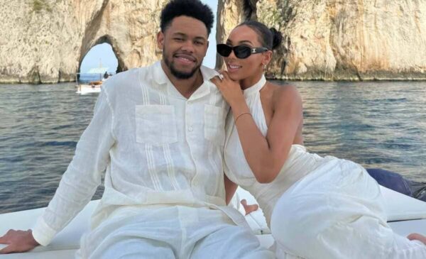 Couple in white outfits sit close on a boat, smiling at the camera with a rocky arch and blue water in the background.