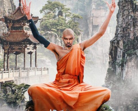 Monk in an orange robe sits cross‑legged on a rocky ledge, arms raised in meditation, with a misty canyon and pagoda behind him.