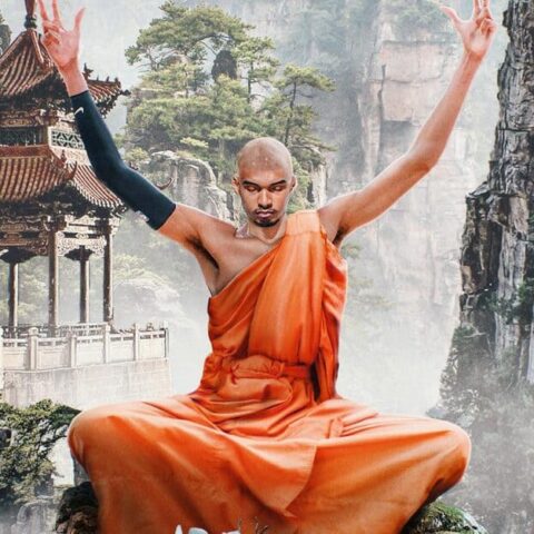 Monk in an orange robe sits cross‑legged on a rocky ledge, arms raised in meditation, with a misty canyon and pagoda behind him.