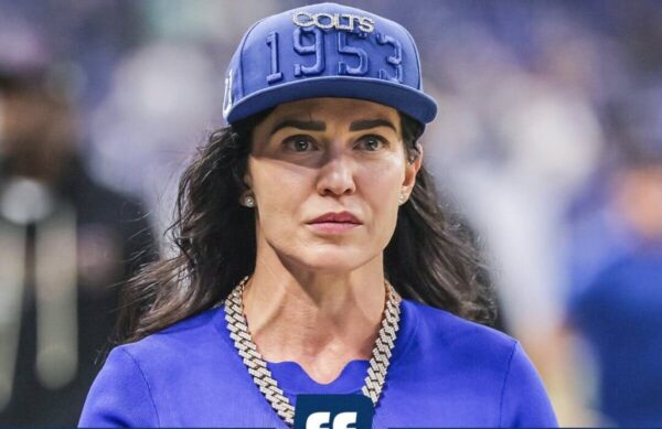 Woman wearing a blue cap embroidered with '1953' and a matching blue top, with a chunky silver chain necklace, looking off to the side.