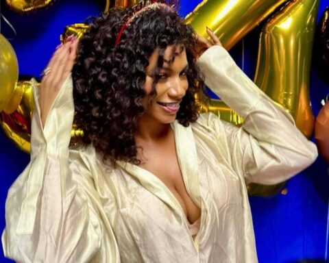 Woman with curly hair in a cream satin blouse posing at a party with gold and pastel balloons in the background.