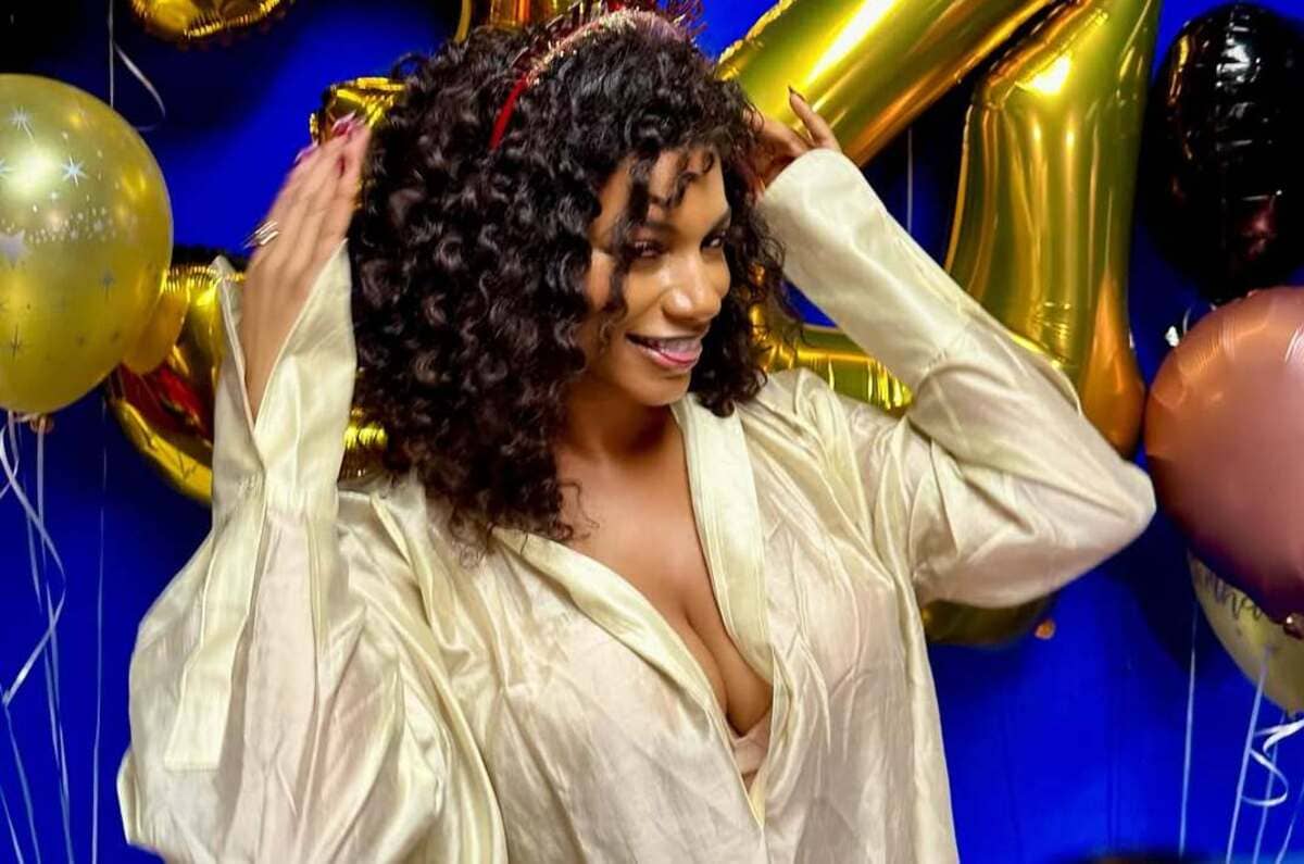 Woman with curly hair in a cream satin blouse posing at a party with gold and pastel balloons in the background.