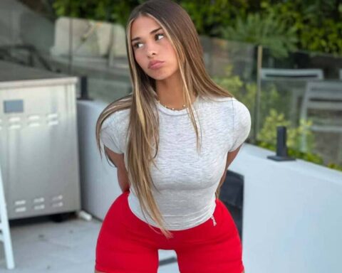 Young woman with long blond hair wearing a gray t-shirt and red shorts, standing outdoors on a patio and looking to the side.