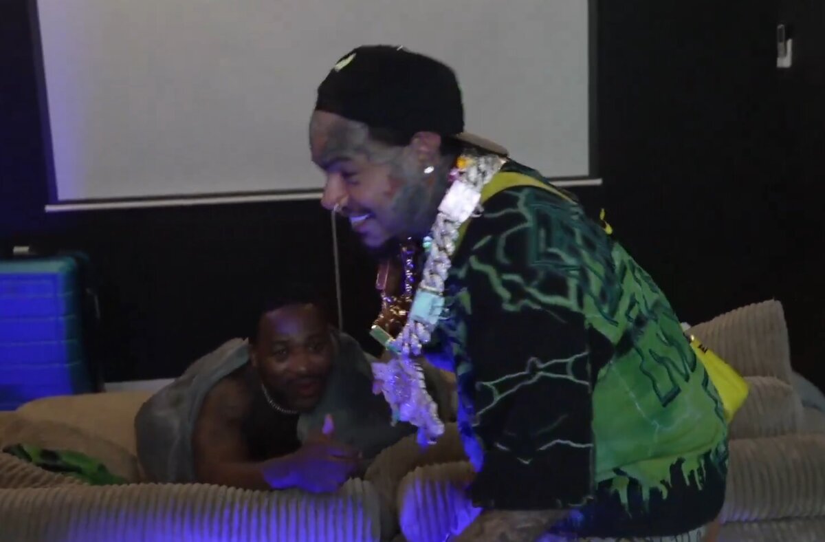 Man in a cap and heavy gold chains leans over a beige couch, smiling at a friend under a blanket in a dim, purple-lit room.