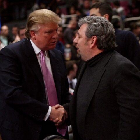 Two men in dark jackets shake hands in a crowded event, one with a pink tie and light hair.