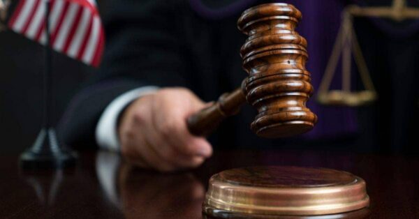 Judicial gavel raised over a sound block in a courtroom, with the scales of justice and an American flag in the background.