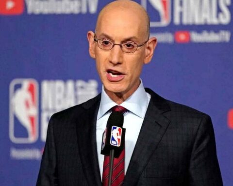 Bald man in a suit speaks at a podium with an NBA logo microphone at a blue press backdrop