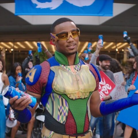 A man in a colorful sci‑fi armor costume and yellow goggles poses with a blue beverage can as a cheering crowd behind him holds up blue cans.