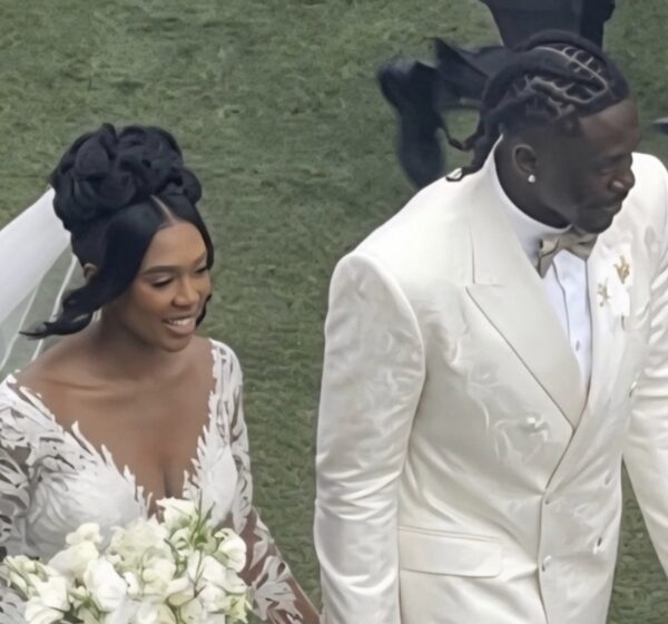Bride in a white lace wedding gown holding a bouquet of white flowers walks beside a groom in a white suit on grass.