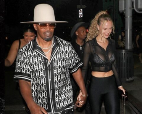 Man in a wide white hat and patterned black‑and‑white shirt walks hand‑in‑hand with a woman in a sheer black top and black leggings at night.