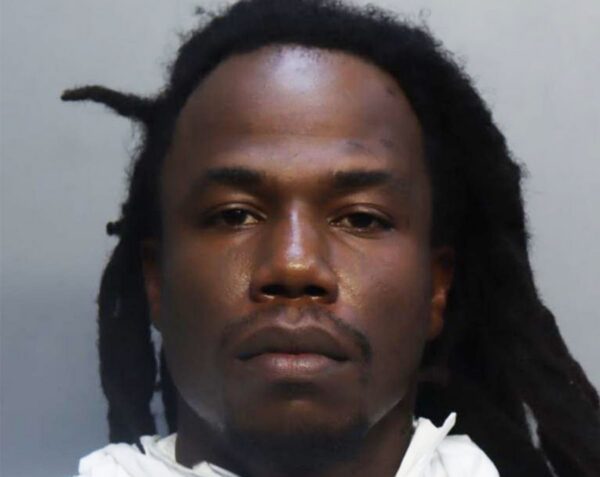 Mugshot of a Black man with dreadlocks, wearing a white jumpsuit, looking slightly left against a gray backdrop.
