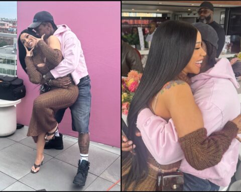 Couple hugs in front of a pink backdrop; woman in brown knit dress, man in a pink hoodie and cap.