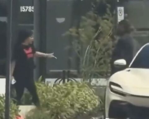Person wearing a mask and black outfit with a pink top points toward a white car parked on the sidewalk in front of a storefront.