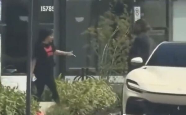 Person wearing a mask and black outfit with a pink top points toward a white car parked on the sidewalk in front of a storefront.