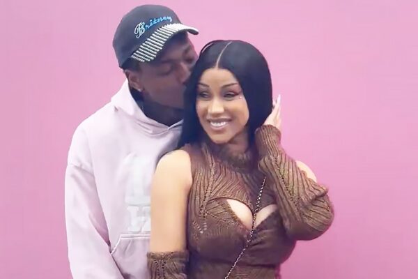 A couple stands against a pink wall; man kisses the woman's temple as she smiles at the camera.