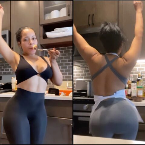 Woman in a black sports bra and leggings raises one arm while eating with a fork in a modern kitchen.