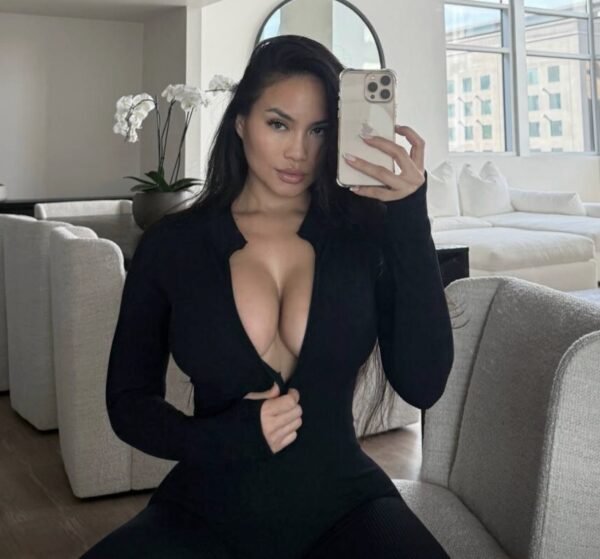 Woman with long dark hair wearing a black plunging top sits on a light sofa, taking a mirror selfie in a bright living room.