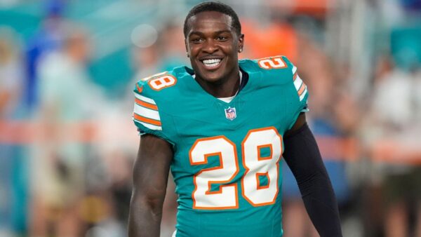 A smiling football player in a teal Miami Dolphins jersey with the number 28 on the chest, stadium crowd blurred in the background.