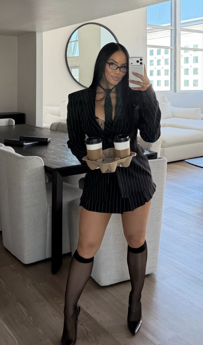 Woman in a pinstripe blazer dress takes a selfie, holding a tray with two coffee cups in a bright living room.