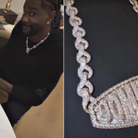 Smiling man in a black outfit shows off a sparkling diamond necklace while sitting at a table with a gift box nearby.