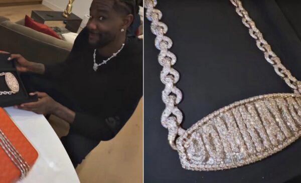 Smiling man in a black outfit shows off a sparkling diamond necklace while sitting at a table with a gift box nearby.