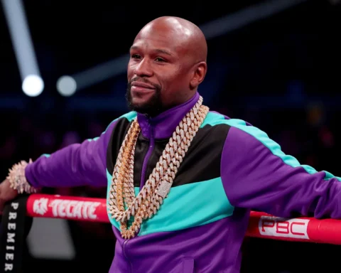 Bald man in a purple and teal tracksuit leaning on boxing ring ropes, wearing multiple chunky gold chains in a crowded arena.