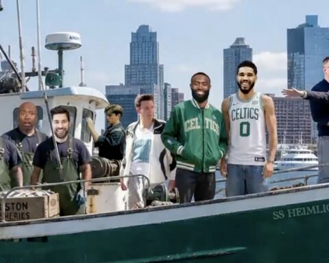 Group of men on a green fishing boat in a harbor, with a city skyline in the background.